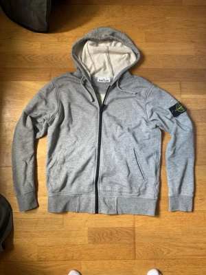 Stone Island zip hoodie  - Säljer en grå hoodie från Stone Island med dragkedja och klassisk logga på vänster ärm. Hoodien har två fickor framtill och är tillverkad i mjukt bomullsmaterial. Perfekt för en avslappnad och stilren look.