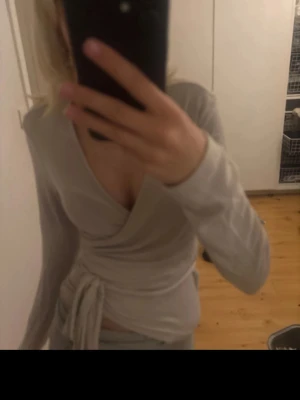 Beige omlottopp med knytband - Säljer en beige omlottopp med långa ärmar och knytband i midjan. Toppen har djup v-ringning och är i ett mjukt, stretchigt material som sitter snyggt på kroppen