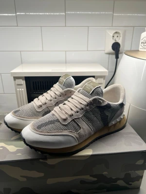 Valentino grå sneakers med camo - Snygga sneakers från Valentino i grått och beige med camo-detaljer på sidan. Ovandelen är i mesh och mocka med vita och ljusgrå partier. Skorna har vita snören, gummisula och coola nitar på hälen. Perfekta för dig som gillar streetstyle och exklusiva märken.