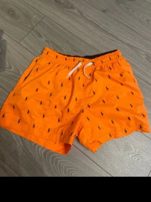 Orange badbyxor från Polo Ralph Lauren - Snygga orange badbyxor från Polo Ralph Lauren med klassiska broderade loggor i svart över hela tyget. Modellen har resårmidja med vit dragsko och sidofickor. Perfekta för stranden eller poolen och ger en färgstark look.
