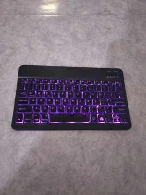 Svart trådlöst tangentbord med bakgrundsbelysning - Säljer ett kompakt svart trådlöst tangentbord med snygg lila bakgrundsbelysning. Tangentbordet har låg profil, rundade hörn och smidig design. Perfekt för surfplatta eller laptop. Materialet är plast och det finns reglage för att slå på/av samt ansluta.