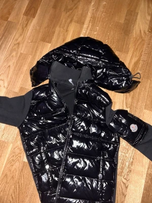 Svart glansig dunväst från Moncler - Snygg svart dunväst från Moncler med glansigt material och avtagbar huva. Västen har dragkedja framtill, ribbade svarta ärmar och Moncler-logga på armen. Insidan har ett unikt serietryck. Perfekt för dig som vill ha en trendig och varm look.
