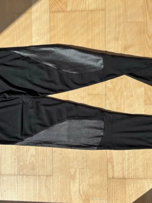 Svarta leggings med glansiga detaljer - Säljer ett par svarta leggings med glansiga, nästan metallic-liknande paneler längs låren och vaderna. Tight passform och stretchigt material som ger en sportig look. 