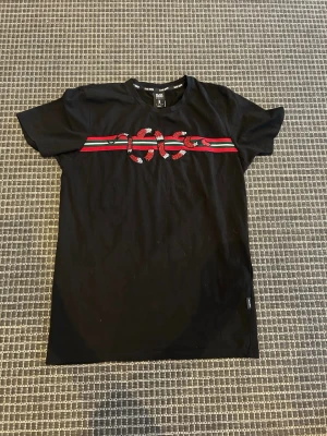 Svart t-shirt med ormtryck Black Squad - Svart t-shirt från Black Squad med slim fit-passform. Framtill finns ett tryck med en orm över röda och gröna ränder. Perfekt för dig som gillar streetwear och vill sticka ut med en cool detalj. Klassisk rund hals och korta ärmar.