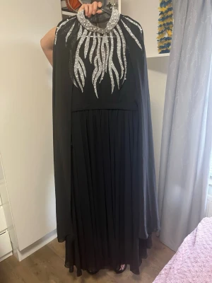 Svart långklänning med silverdetaljer - Elegant svart långklänning med glittriga silverapplikationer runt halsen och på ärmen. Klänningen har en cape-liknande överdel i tunt tyg och ett flytande fall. Perfekt för speciella tillfällen där du vill sticka ut. Jag köpte detta på Instagram (@lerich.se) 