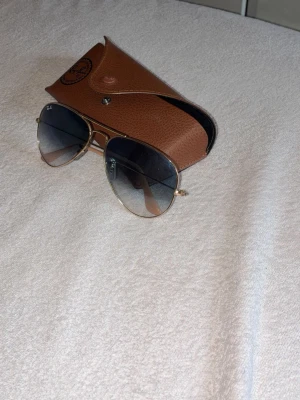 Ray-Ban solglasögon guld - Säljer ett par klassiska Ray-Ban aviator solglasögon med guldfärgad metallbåge och tonade blå-svarta glas. Kommer med original brunt fodral i skinnimitation. Perfekt accessoar för att höja din stil i solen.