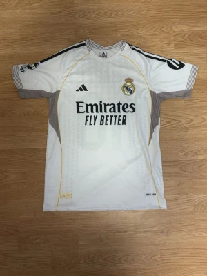 Real Madrid matchtröja Adidas vit - Snygg vit Real Madrid matchtröja från Adidas med klubbmärke och sponsortryck på bröstet. Tröjan har svarta och gula detaljer, samt grå meshpaneler på sidorna för ventilation. Perfekt för fotbollsträning eller att bära till vardags.