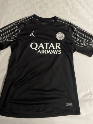Svart PSG x Jordan matchtröja - Snygg svart matchtröja från PSG i samarbete med Jordan. Tröjan har vita detaljer, PSG-logga och Jordan-logga på bröstet samt Qatar Airways-tryck. Unik design med vita linjer på axlarna. Perfekt för fotboll eller streetwear.