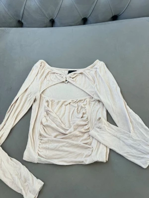 Beige långärmad topp från SHEIN - Trendig beige långärmad topp från SHEIN med cut-out detalj framtill och dekorativ knut med pärlor vid halsen. Toppen har rynkade detaljer på magen och är tillverkad i ett mjukt, stretchigt material. Perfekt för dig som gillar unika detaljer och en modern look.
