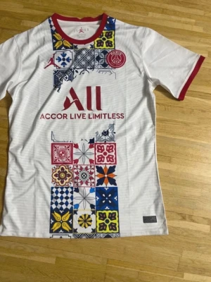 PSG x Jordan vit t-shirt med mönster - Säljer en vit PSG x Jordan t-shirt med färgglada patchwork-mönster framtill och röda detaljer vid krage och ärmslut. T-shirten har tryckt PSG-logga, Jordan-logga och texten 'Accor Live Limitless' på bröstet. Perfekt för dig som gillar sportig och unik stil.