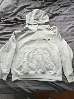 Grå Essentials Fear of God hoodie - Snygg ljusgrå hoodie från Essentials Fear of God med svart logga på bröstet och stor text på ryggen. Hoodien har en stor känguruficka framtill, ribbade muddar och en rejäl huva. Perfekt för dig som gillar streetwear och vill ha en clean look.