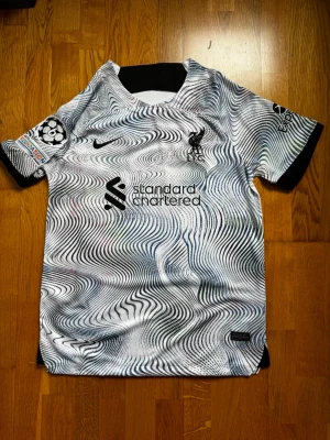 Liverpool bortatröja Nike svartvit - Säljer en Liverpool FC bortatröja från Nike med svartvitt psykedeliskt mönster och svarta detaljer. Tröjan har korta ärmar, klubbmärke och sponsortryck på bröstet samt Champions League-märke på ärmen. Tillverkad i lätt och ventilerande material.