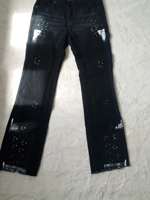 Svarta flared jeans size 30 - Säljer ett par svarta jeans med raka ben och råa benslut. Jeansen har vita färgstänk och slitna detaljer framtill för en edgy look. Perfekta för dig som gillar streetstyle och vill sticka ut lite extra.