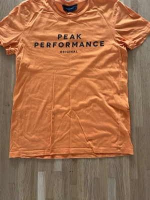 Peak Performance t-shirt - Snygg orange t-shirt från Peak Performance med svart tryck framtill. Klassisk rund hals och korta ärmar. Tillverkad i mjuk bomull som känns skön mot huden. Perfekt för dig som gillar stilrena och sportiga plagg.