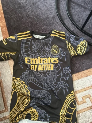 Real Madrid Bellingham tröja drake - Säljer en svart Real Madrid fotbollströja med gult drakmönster och gula detaljer. På framsidan står det 'Emirates Fly Better' och klubbmärket, på baksidan står det 'Bellingham' och nummer 5. Korta ärmar och rund hals. Perfekt för dig som älskar fotboll och snygg design.