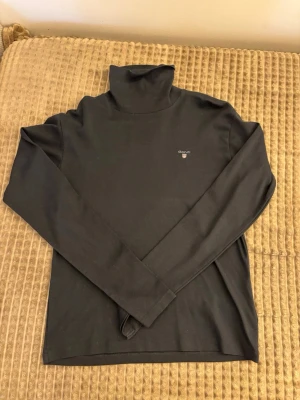 Svart polotopp från Gant - Svart polotopp från Gant med diskret logga på bröstet. Topp i mjukt material med lång ärm och klassisk polokrage. Perfekt att styla lager på lager eller som den är för en clean look.