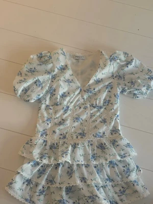 Blommig kortklänning med volanger - Supersöt kortklänning från  Design By Amor i vitt med blått blommönster. Klänningen har puffärmar, v-ringning, spetsdetaljer och flera lager volanger. Perfekt för sommarens alla tillfällen!
