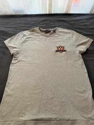 Grå t-shirt från GANT med brodyr - Snygg grå t-shirt från GANT med ett broderat emblem på bröstet i marinblått, rött och vitt. Klassisk rund halsringning och korta ärmar. Tillverkad i mjuk bomull för en skön känsla. Perfekt till jeans eller shorts.