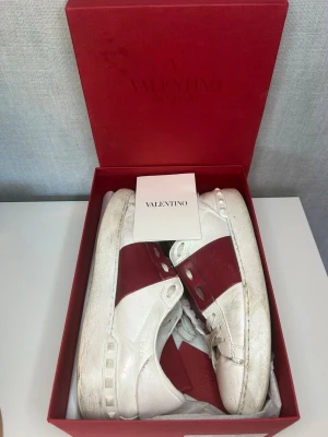 Röda valentino opens  - Snygga vita sneakers från Valentino med en bred röd läderdetalj över snörningen och nitar på hälen. Skorna är låga och har en chunky sula. Tillverkade i läder och har klassisk logga på plösen. Perfekta för dig som vill sticka ut med exklusiv design. Allt på bilden medföljs 