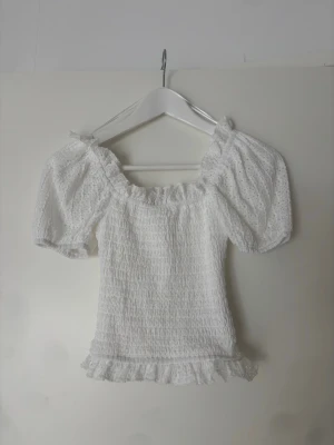 Vit blus med puffärm och volang - Söt vit blus med puffiga kortärmar och smockad kropp. Blusen har volangkant nedtill och runt ärmsluten samt broderade detaljer i tyget. 