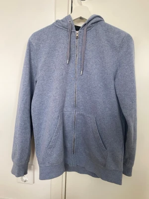 Ljusblå zip hoodie med huva - En enkel och stilren ljusblå hoodie med dragkedja framtill och stor känguruficka. Hoodien har justerbar huva med snören och ribbade muddar vid ärmar och nederkant. Perfekt för chill dagar eller när du vill ha något bekvämt. Materialet känns mjukt och skönt mot huden. Liten fläck (se bild)