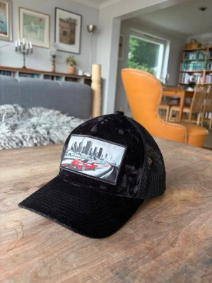 Exklusiv Revenite keps - (LIMITED EDITION) Säljer en svart keps med mesh på sidorna och ett stort tygmärke framtill som visar en röd sportbil mot en stadssilhuett. Kepsen har böjd skärm och justerbar snapback baktill. Perfekt för dig som gillar streetstyle och coola detaljer.