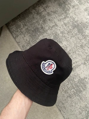Moncler hatt  - Snygg svart bucket hat från Moncler med det klassiska broderade logotypmärket framtill i vitt, blått och rött. Hatten är tillverkad i ett mjukt tygmaterial och har en avslappnad passform som ger en cool och trendig look. Inga skador eller hål!