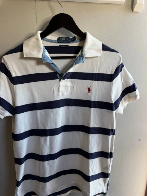 Randig pikétröja från Polo Ralph Lauren - Klassisk vit pikétröja med mörkblå breda ränder från Polo Ralph Lauren. Tröjan har korta ärmar, krage och knappar vid halsen samt den ikoniska röda broderade loggan på bröstet. Tillverkad i bomull för en skön och luftig känsla.