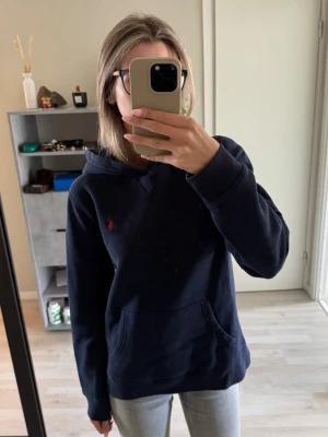 Mörkblå hoodie från Ralph Lauren - Säljer en mörkblå hoodie från Ralph Lauren med klassisk huva och känguruficka. Tröjan har det ikoniska röda Polo-logotypen broderad på bröstet. Tillverkad i mjukt bomullsmaterial och har ribbade muddar vid ärmslut och nederkant.