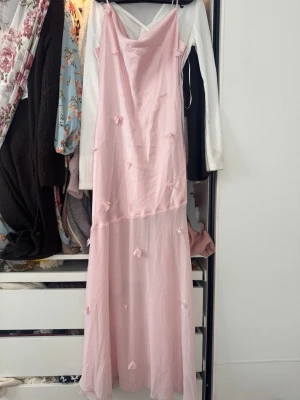 Gina Tricot Lång ljusrosa klänning - Söt långklänning i ljusrosa med tunna axelband och små dekorativa rosetter. Klänningen har en lätt transparent nederdel och är perfekt för sommarens tillfällen. Materialet känns tunt och luftigt. Passar för balen, dock blev klänningen trasig på sidan och är sydd över, högra lägre bröst