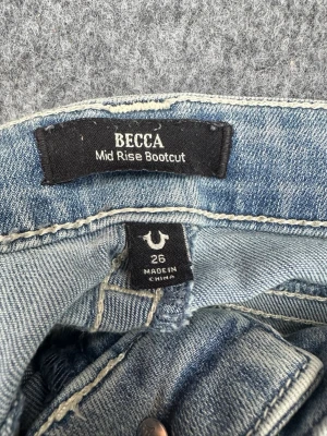 True Religion Becca Bootcut Jeans - Snygga ljusblå jeans från True Religion, modell Becca Mid Rise Bootcut. Klassisk femficksdesign med dekorativa sömmar och knappstängning på bakfickan. Perfekt för dig som gillar en avslappnad men trendig look. Jättefina men lite slitna vid hälen.