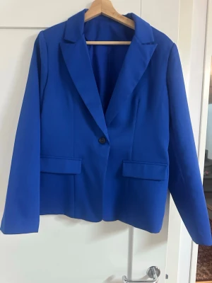 Kornblå kavaj med klassisk passform - Stilren kornblå kavaj med en knapp framtill och två fickor med lock. Klassisk krage och rak passform, perfekt för att lyfta vilken outfit som helst. Materialet känns slätt och har en lätt struktur. Snygg att matcha med jeans eller kostymbyxor.