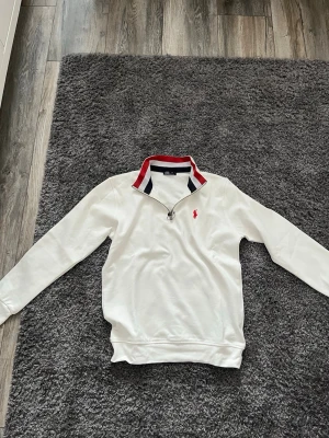 Vit quarter zip från Ralph Lauren - Snygg vit quarter zip tröja från Ralph Lauren med röd broderad logga på bröstet. den är inte använd min pappa köpte den fast den satt för stor på mig därför söker jag till att sälja den riktigt bra skick ungefär helt ny. Pris kan diskuteras 