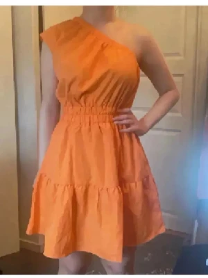 Orange/koral oneshoulder kortklänning - Snygg orange/koral kortklänning med oneshoulder-design och resår i midjan. Klänningen har volangdetaljer och är tillverkad i ett lätt bomullsliknande material.  Mer åt det korala färgen men svårt att få på bild❤️