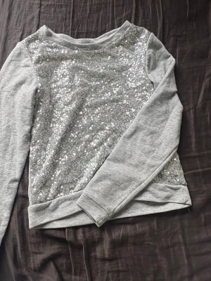 Grå sweatshirt med paljetter - Supersnygg grå sweatshirt med glittriga silverpaljetter på framsidan. Tröjan har rund halsringning och långa ärmar i mjukt sweatshirtmaterial. Perfekt för dig som vill sticka ut lite extra med en touch av glitter.