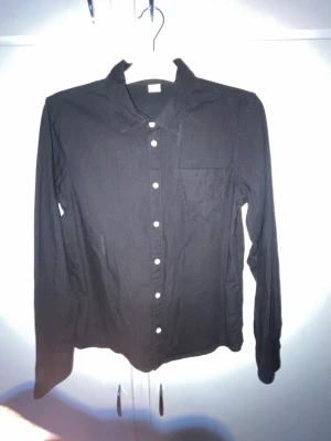 Svart skjorta med vita knappar - Klassisk svart skjorta med långa ärmar och vita knappar framtill. Skjortan har en bröstficka och enkel krage. Perfekt att styla till jeans eller kjol för en clean look.