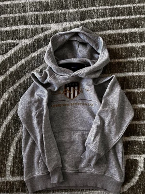Grå hoodie från GANT med tryck - Snygg grå hoodie från GANT med huva och känguruficka. På bröstet finns ett broderat emblem i rött, vitt och guld samt texten 'American Sportswear'. Tillverkad i mjukt bomullsmaterial, perfekt för en avslappnad och sportig stil.