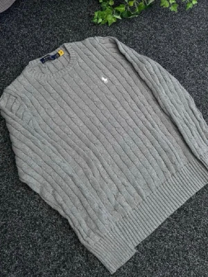 Grå kabelstickad tröja Polo Ralph Lauren - Snygg grå kabelstickad tröja från Polo Ralph Lauren med rund halsringning och klassisk vit broderad logga på bröstet. Tröjan har ribbade muddar vid ärmslut och nederkant, perfekt för en stilren och avslappnad look Det står den är storlek M men den passar bäst som S