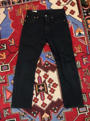 Svarta Levi's 501 jeans straight fit - Klassiska svarta Levi's 501 jeans med rak passform och fem fickor. Jeansen har knappgylf, nitar i kopparfärg och Levi's ikoniska läderpatch bak i midjan. Tillverkade i kraftig denim, perfekta för en tidlös och avslappnad look.