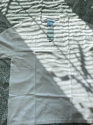 Vit NOCTA x Nike t-shirt - Säljer en clean vit t-shirt från Nike och NOCTA-kollektionen. T-shirten har korta ärmar, rund hals och tryckt NOCTA-logga i svart på ryggen samt liten Nike Swoosh på bröstet. Tillverkad i mjuk bomull för skön passform.