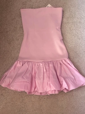 Ljusrosa axelbandslös klänning Zara - Supersöt ljusrosa axelbandslös klänning från Zara med volangig kjol och figurnära passform upptill. Klänningen har en kort längd och är perfekt för dig som vill sticka ut med en feminin och trendig look. Materialet känns mjukt och har en lätt glans.