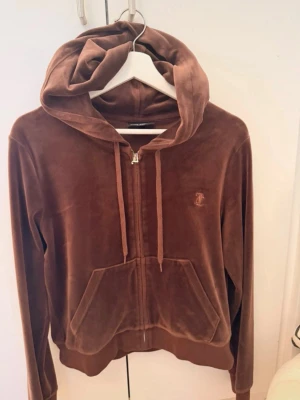 Brun velour hoodie med dragkedja - Säljer en brun velour hoodie från Juicy Couture med dragkedja framtill och känguruficka. Hoodien har en broderad logga på bröstet och justerbar huva med snören. Perfekt för en chill och trendig look.