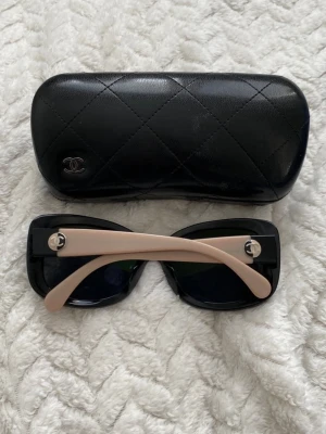 Chanel beige och svarta solglasögon - Säljer ett par ikoniska Chanel solglasögon i beige och svart acetat med polariserade linser. Modellen har en fyrkantig form och klassiska Chanel-loggor på skalmarna. Kommer med ett quiltat svart fodral i skinn. Perfekt accessoar för en trendig look.