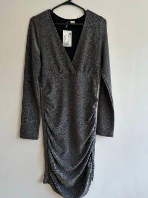 Glittrig silverklänning från H&M - Snygg silverfärgad klänning från H&M med glittrigt material, djup V-ringning och långa ärmar. Klänningen har rynkade detaljer längs sidorna och en kroppsnära passform som ger en trendig look. Perfekt för dig som vill sticka ut.