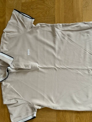 Beige piké från BOSS - Stilren beige pikétröja från BOSS med korta ärmar och svart-vit rand på krage och ärmslut. Tröjan har knäppning framtill och diskret BOSS-logga broderad på bröstet. Perfekt för en clean och klassisk look.
