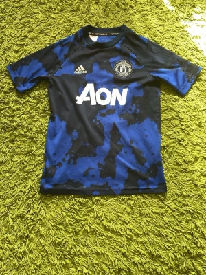 Manchester United träningströja blå/svart - Snygg Manchester United träningströja från Adidas i blått och svart camo-mönster. Kortärmad modell med klubbmärke och Adidas-logga på bröstet samt AON-tryck framtill. Perfekt för fotbollsträning eller gymmet.
