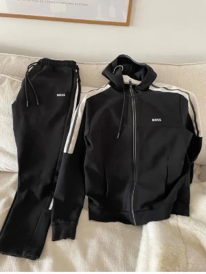 Svart zip hoodie från BOSS - Snygg svart zip hoodie från BOSS med vita ränder längs ärmarna och logga på bröstet. Tröjan har huva med dragsko, ribbade muddar och dragkedja framtill. Perfekt för en sportig och avslappnad stil.