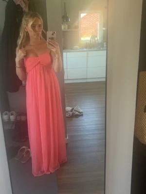 Lång rosa axelbandslös klänning - Säljer en lång, axelbandslös klänning i en härlig rosa/voral färg. Klänningen har ett twistat bröstparti och faller snyggt i ett luftigt, svepande tyg. Perfekt för sommarens alla tillfällen!