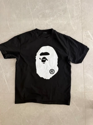 Svart BAPE t-shirt med stort tryck - Svart t-shirt från BAPE med det ikoniska vita apansikte-trycket på både fram- och baksidan. Klassisk passform och rund halsringning. Perfekt för dig som gillar streetwear och vill sticka ut med en tydlig logga.