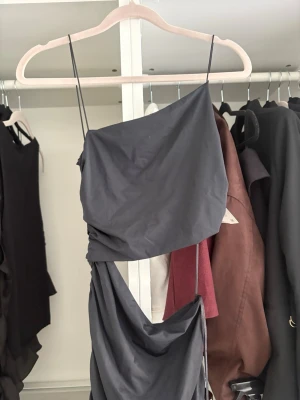 Svart oneshoulder långklänning - Snygg svart långklänning med oneshoulder-design och tunna axelband. Klänningen har draperade detaljer längs hela längden och en asymmetrisk överdel. Cut out i midjan. Prislapp på